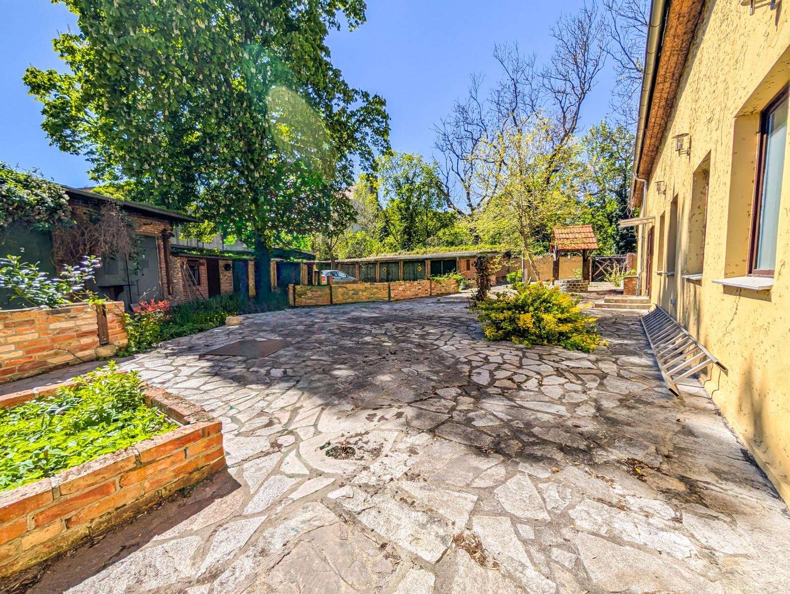 Sunny courtyard with cracked stone paving, brick planters, and a yellow building on the right under a blue sky.