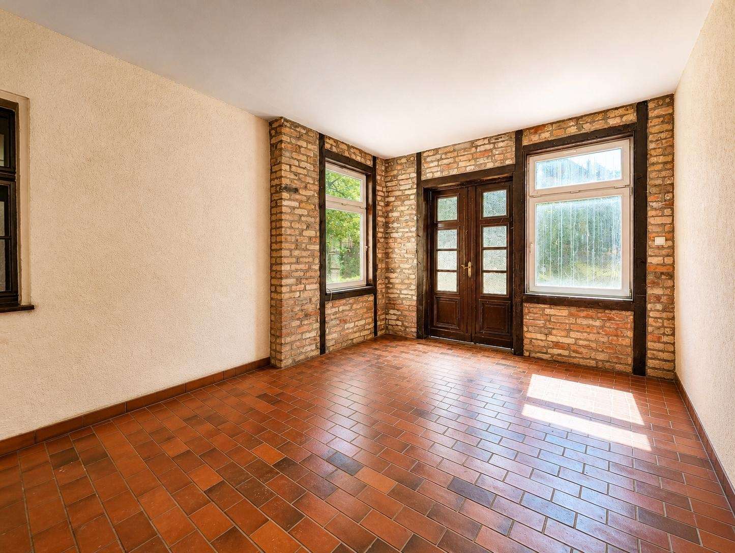 Sunlit room with exposed brick columns, dark wood-framed doors, and large windows over terracotta tile floor.
