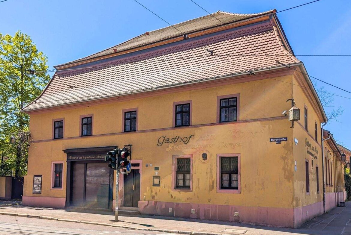 Two-story yellow building with pink trim on a city street, tram tracks in front and overhead wires against a bright blue sky.