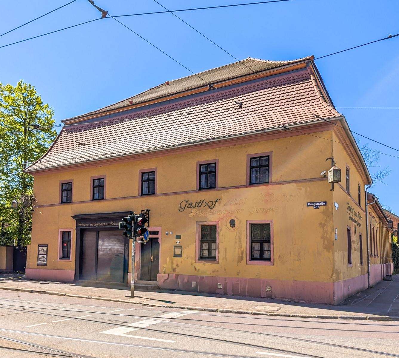 Two-story yellow building with pink trim on a city street, tram tracks in front and overhead wires against a bright blue sky.