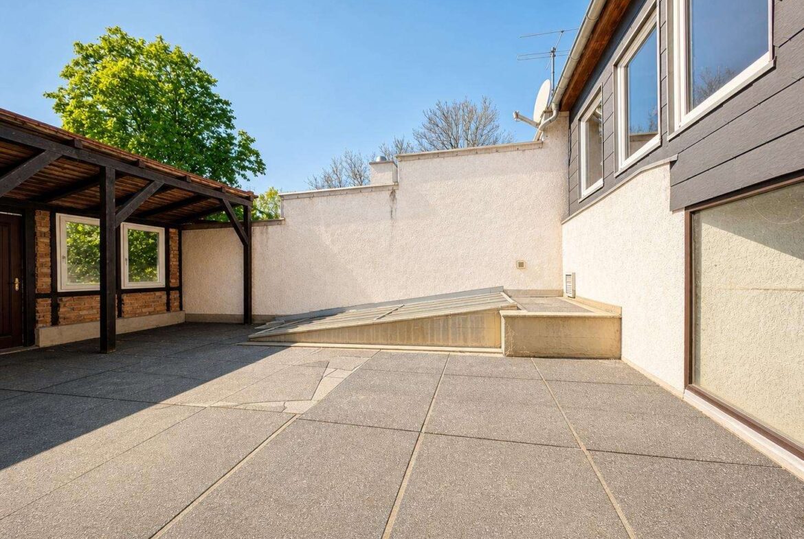 Open-air rooftop terrace with gray concrete tiles, white walls, and a wooden pergola on the left; house windows on the right under a blue sky.