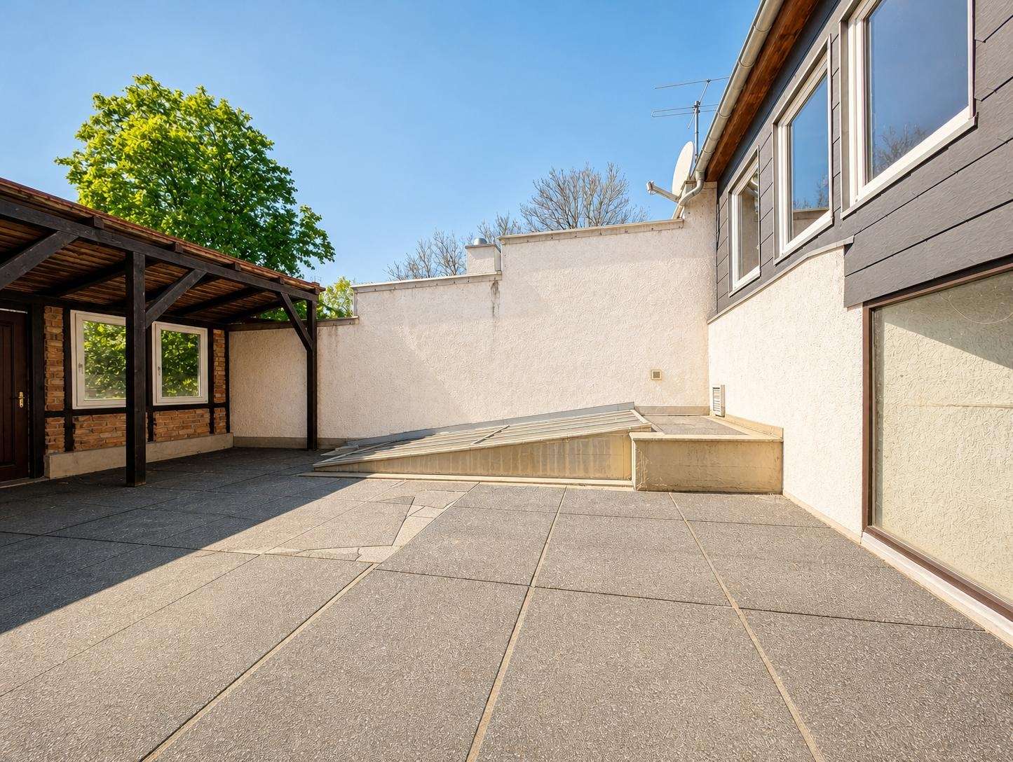 Open-air rooftop terrace with gray concrete tiles, white walls, and a wooden pergola on the left; house windows on the right under a blue sky.