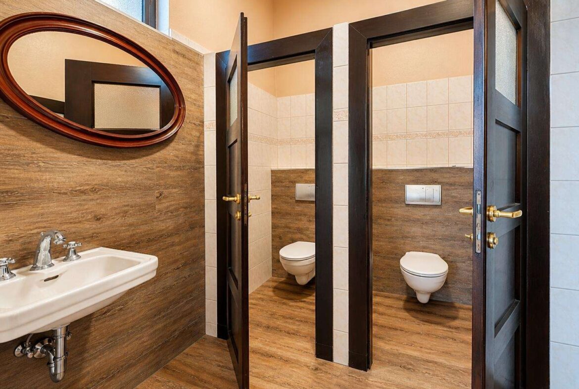 Public restroom with two open stalls and a sink on a wood-paneled wall, a large oval mirror above the sink, and dark doors.
