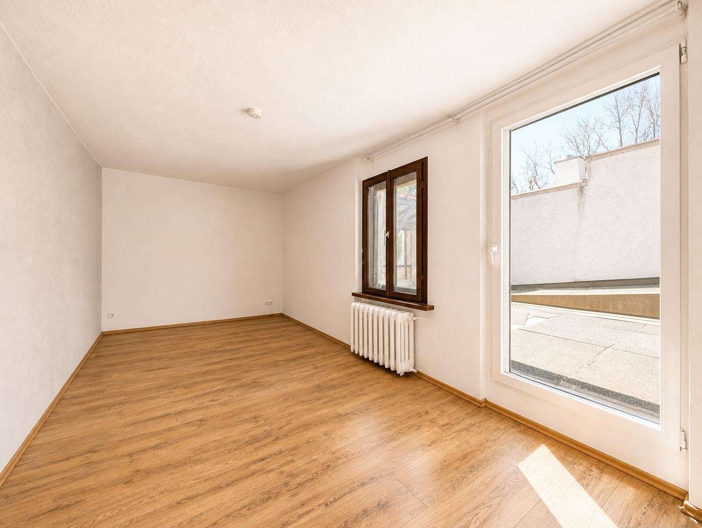 Empty living room with light wood laminate flooring, white walls, a radiator beneath a brown-framed window, and a glass door opening to a sunny patio outside.