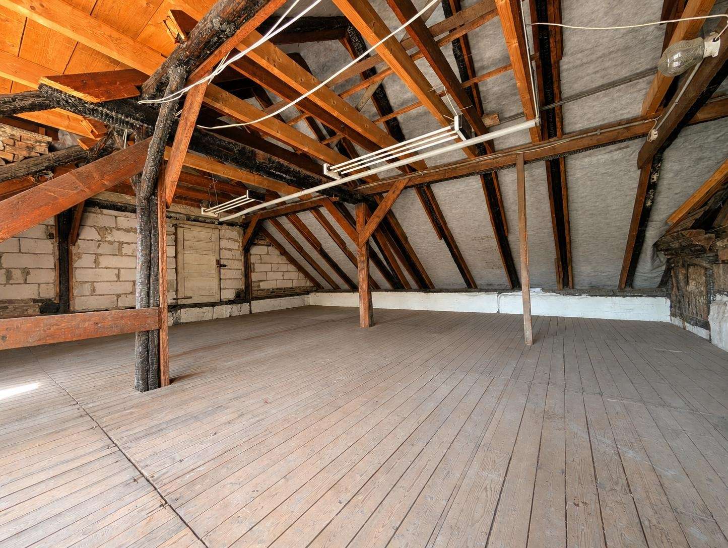 Empty attic space with wooden floorboards and exposed framing, sloped roof, and support beams under insulation-covered walls and ceiling.