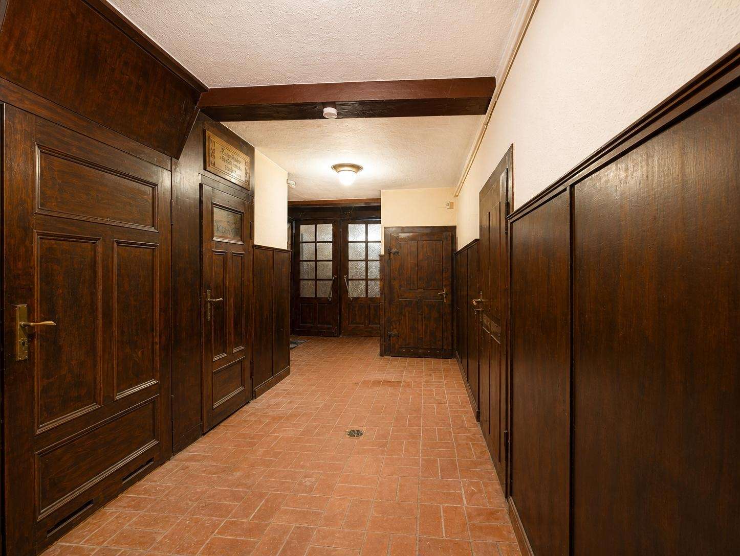 Long hallway lined with dark wood paneled doors and a brick-red tile floor, lit by a central ceiling light.