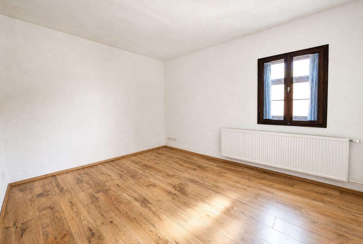 Empty white-walled room with a wooden floor, a radiator along the wall, and a dark-framed window with blue checkered curtains.