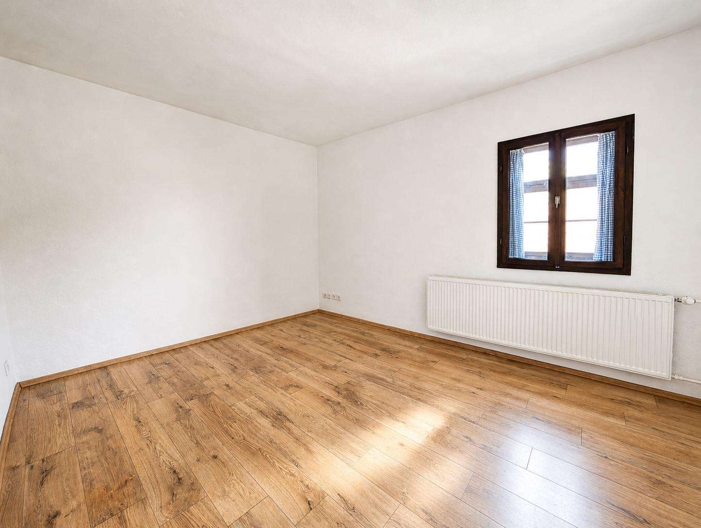 Empty white-walled room with a wooden floor, a radiator along the wall, and a dark-framed window with blue checkered curtains.
