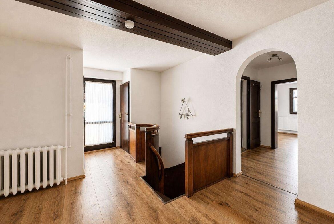Bright interior landing with a dark wood beam ceiling, wooden railing around an open stairwell, and light hardwood floors.