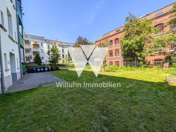 Residential courtyard with green lawn, flanked by white and brick buildings under a clear blue sky; a bicycle and row of trash bins visible along the left edge; watermark 'Willuhn Immobilien' across the center.