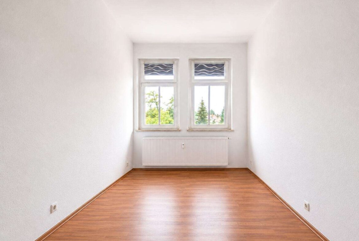 Bright empty room with white walls, two side-by-side windows, radiator below, and a wooden floor.