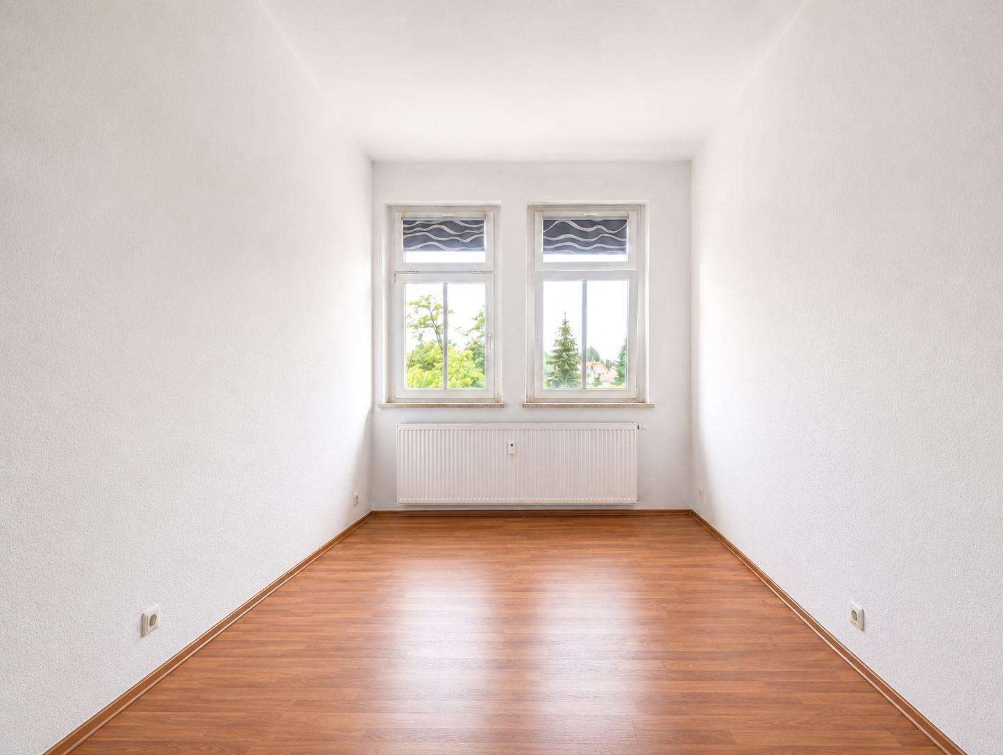 Bright empty room with white walls, two side-by-side windows, radiator below, and a wooden floor.