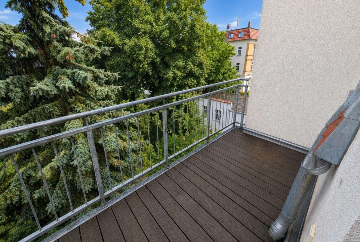 Wooden balcony with metal railing overlooking lush green trees and a neighboring building against a blue sky.