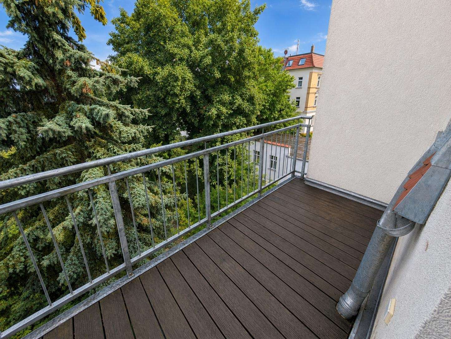 Wooden balcony with metal railing overlooking lush green trees and a neighboring building against a blue sky.