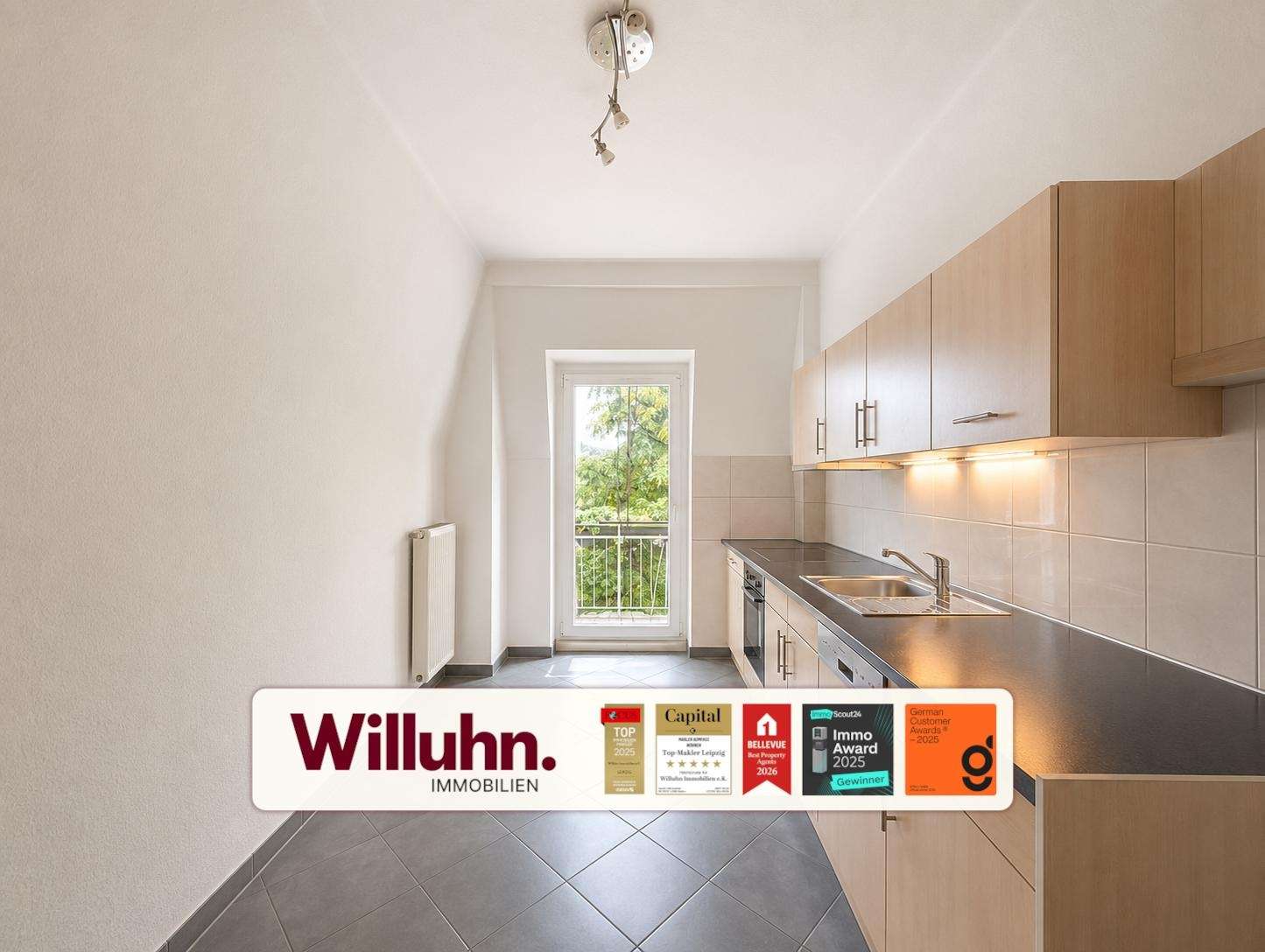 Compact modern kitchen with light wood cabinets, stainless steel counter, sink, and a balcony door; Willuhn Immobilien branding banner along the bottom.