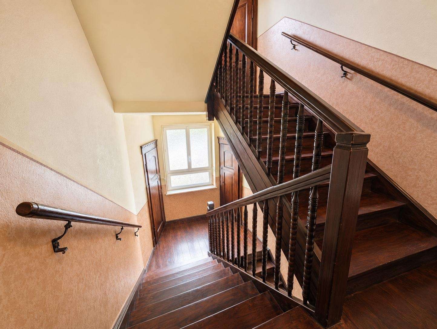 Dark wooden staircase with turned balusters and banister, set against warm beige walls, lit by a window at the landing