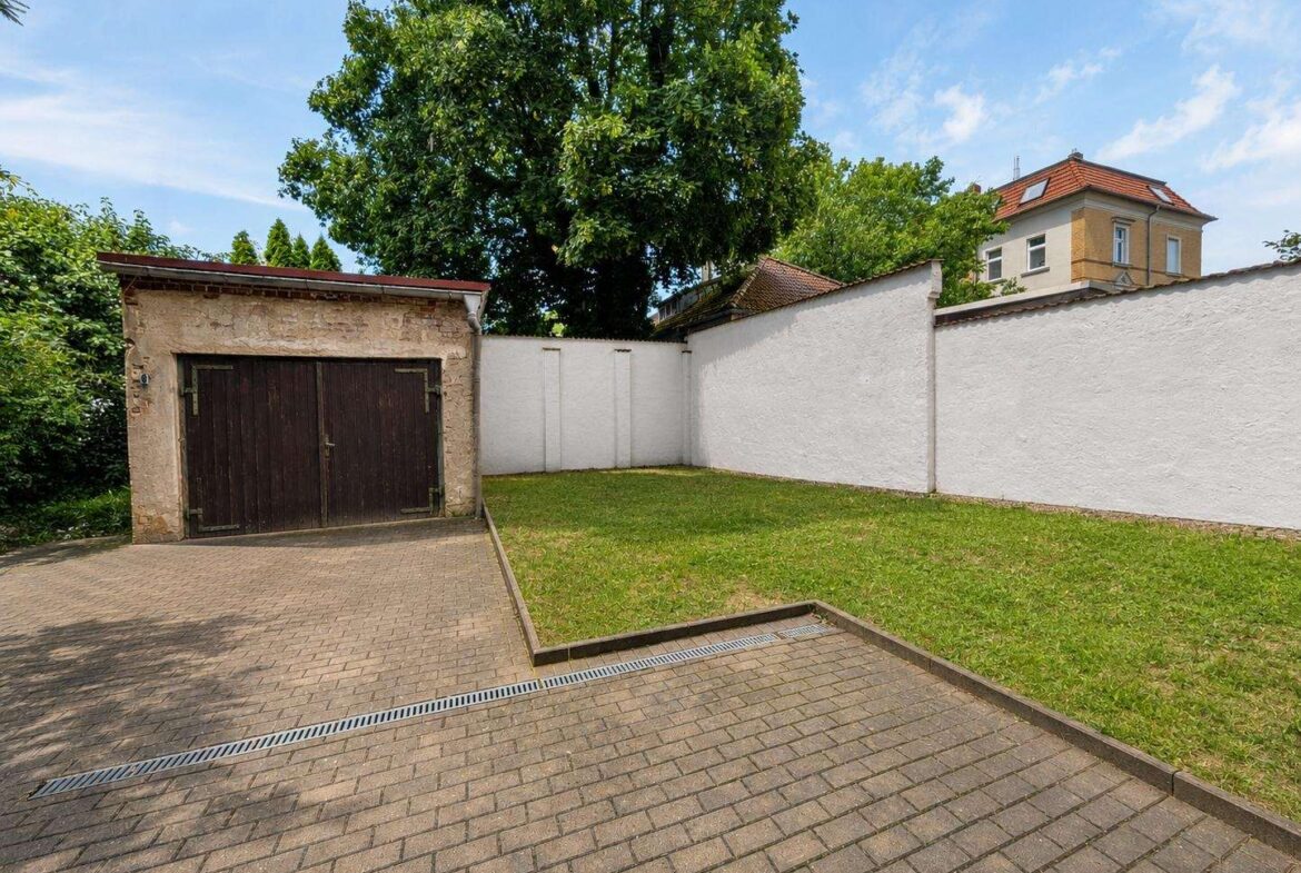 Courtyard with a dark wooden double garage door, white stucco walls, a paved driveway, and a green lawn under a large tree.