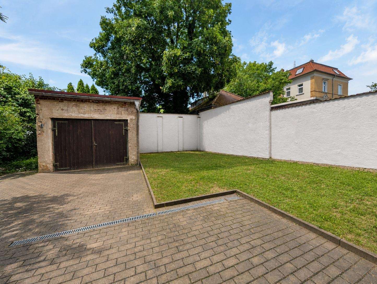 Courtyard with a dark wooden double garage door, white stucco walls, a paved driveway, and a green lawn under a large tree.
