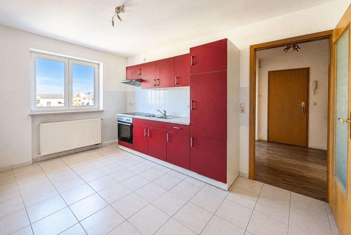 Red kitchen cabinets line a white-tiled wall with a stove, sink, and window view behind them.