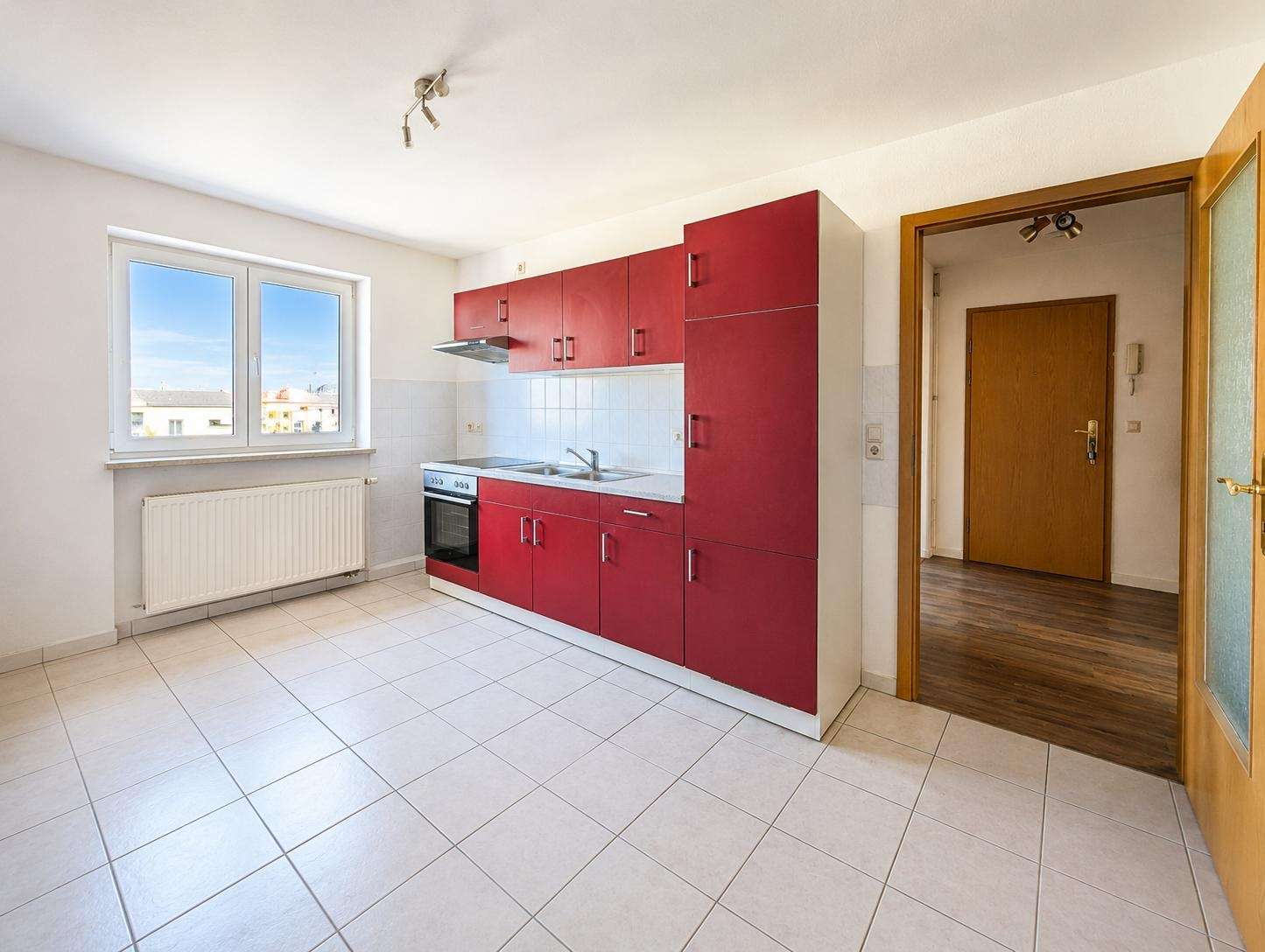 Red kitchen cabinets line a white-tiled wall with a stove, sink, and window view behind them.