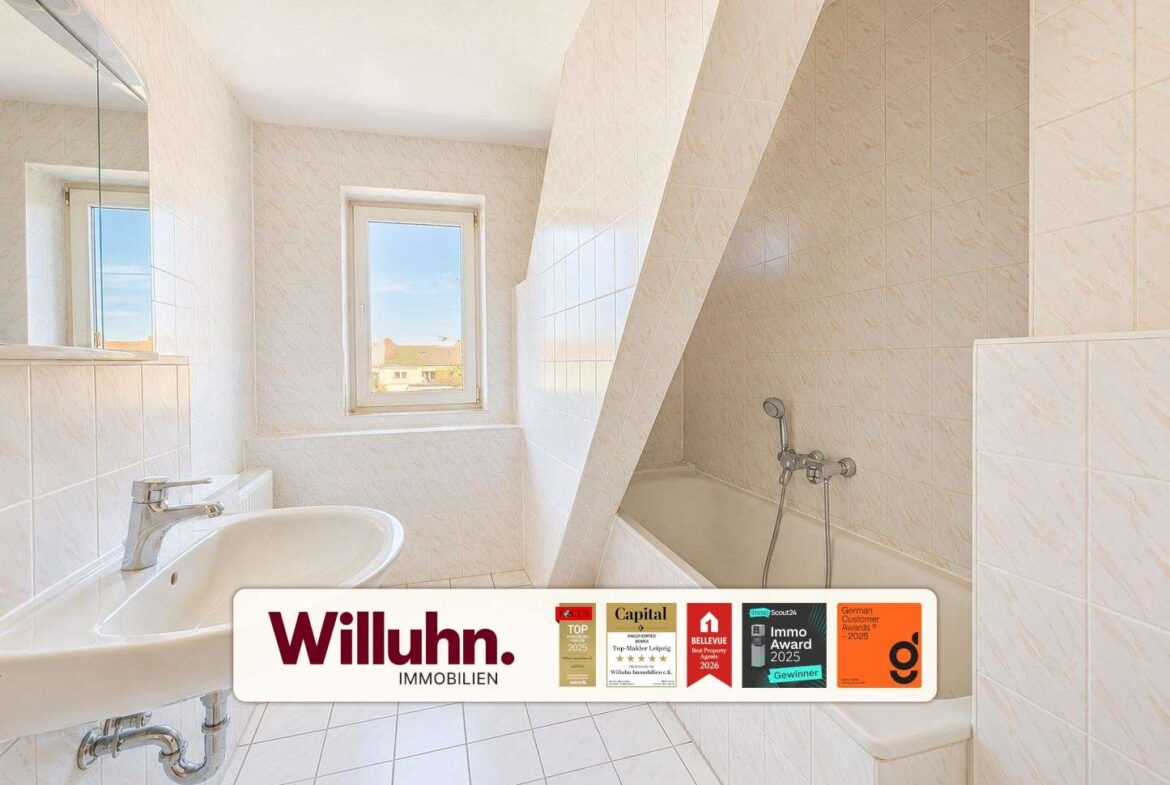 Bright white tiled bathroom with bathtub on the right, sink on the left, and a window letting in sunlight; a slanted wall divides the space; Willuhn Immobilien banner at the bottom.