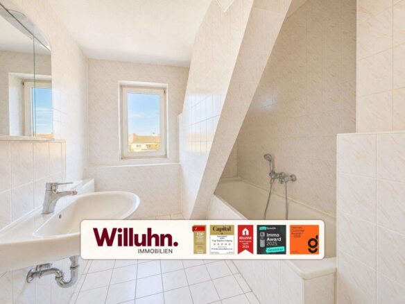 Bright white tiled bathroom with bathtub on the right, sink on the left, and a window letting in sunlight; a slanted wall divides the space; Willuhn Immobilien banner at the bottom.