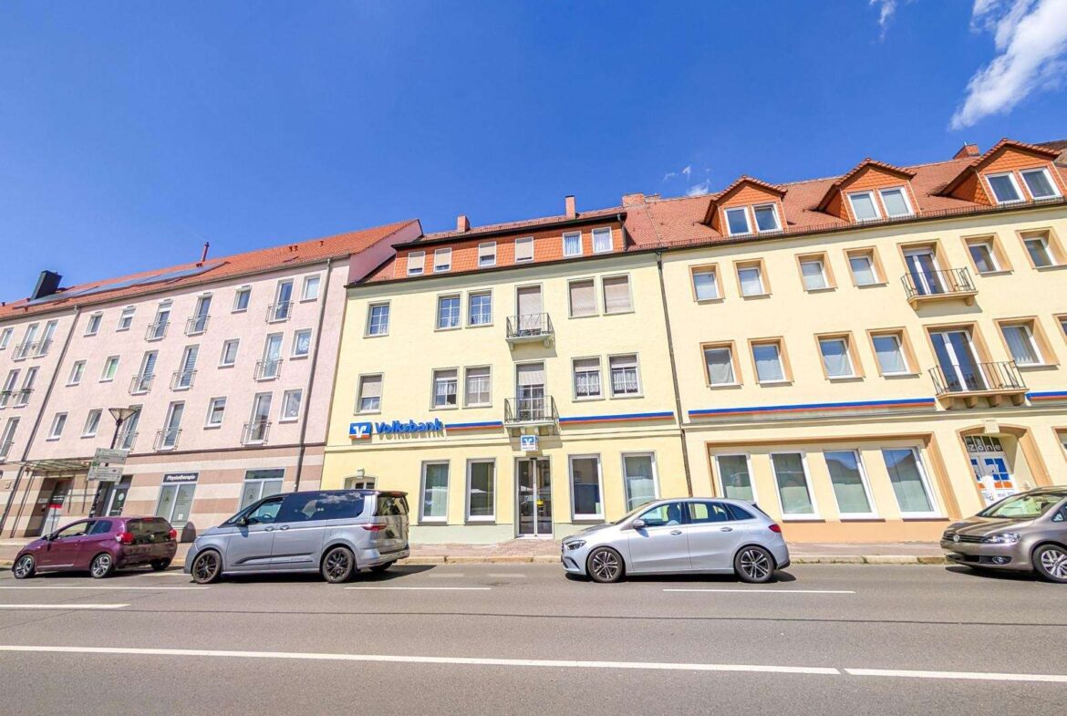 Row of pastel-colored buildings with red-tiled roofs along a sunny street; parked cars in front and a Volkbank sign on the yellow building.
