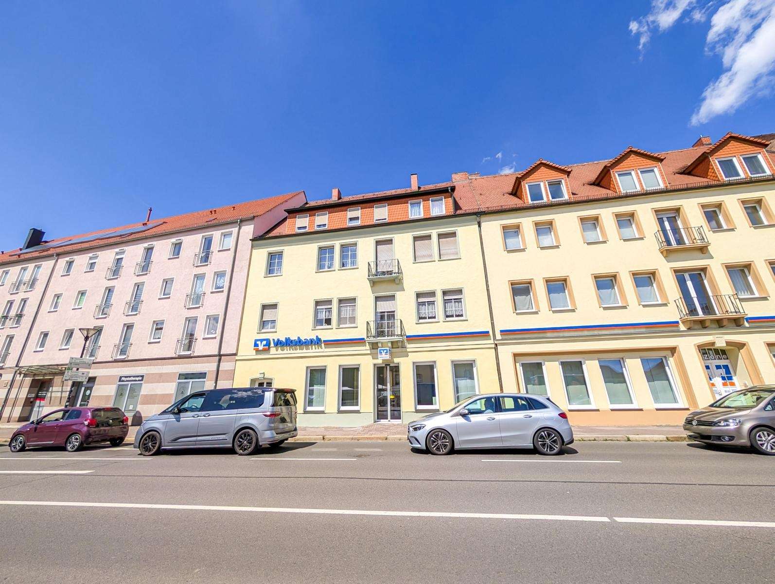 Row of pastel-colored buildings with red-tiled roofs along a sunny street; parked cars in front and a Volkbank sign on the yellow building.