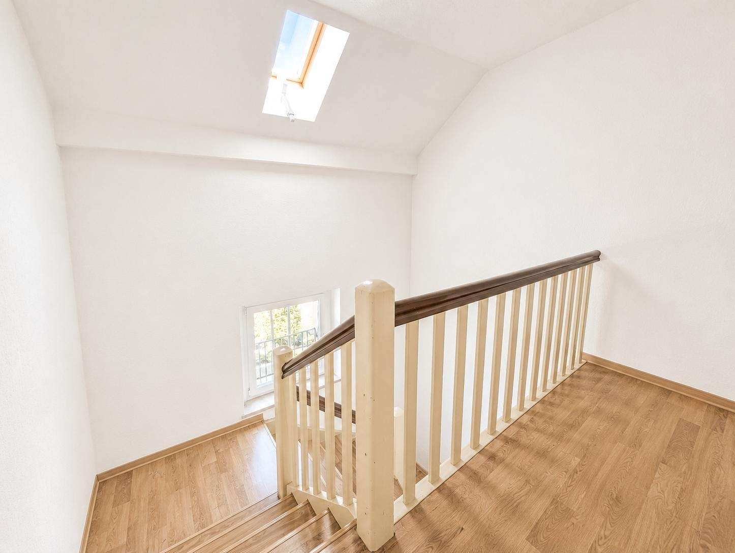 Top of a wooden staircase with a dark handrail, white balusters, and bright white walls; skylight overhead.