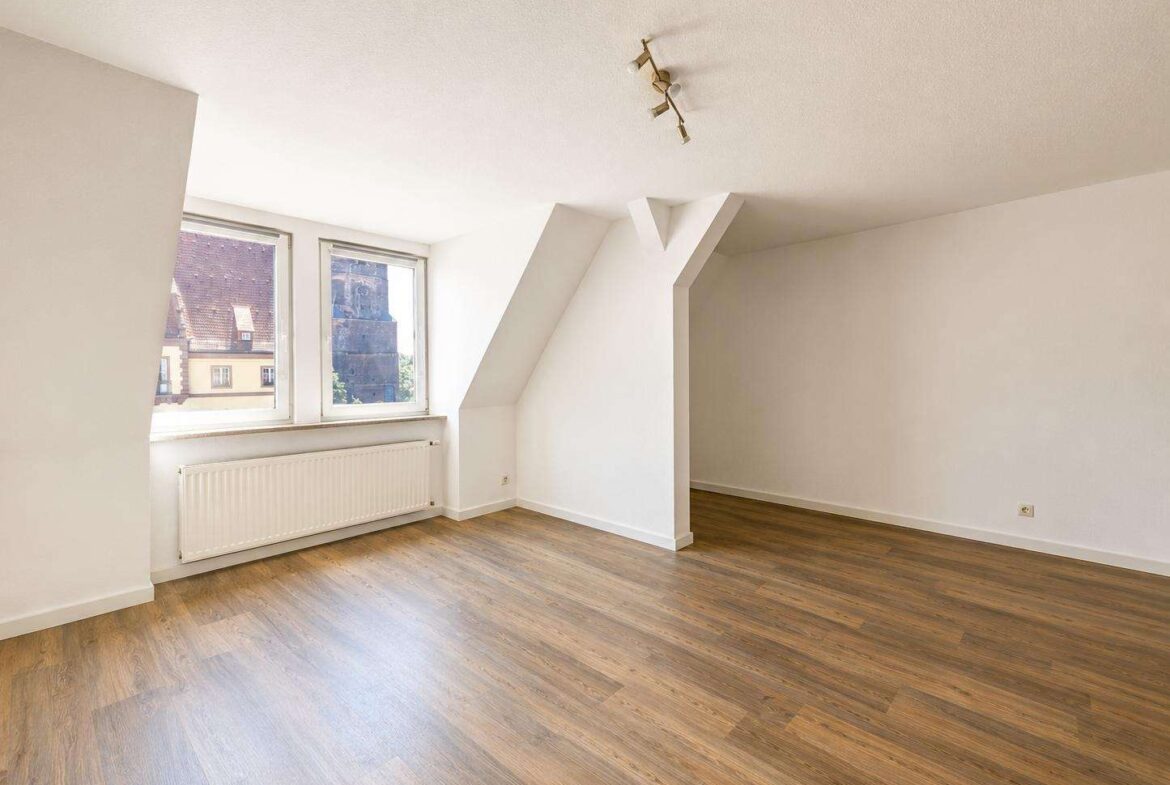Bright empty attic-like room with two large windows, a white radiator, and warm wood floors