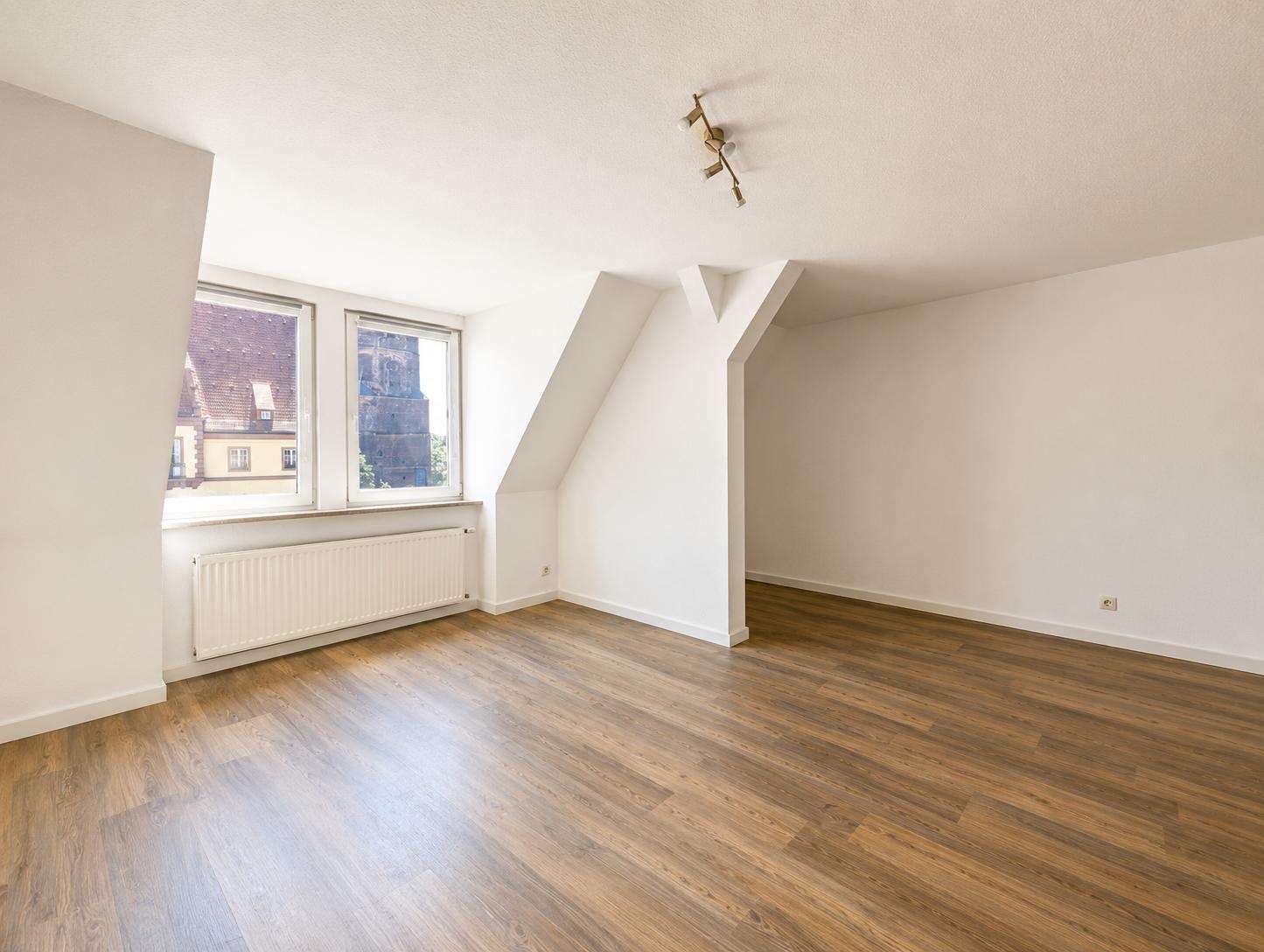 Bright empty attic-like room with two large windows, a white radiator, and warm wood floors