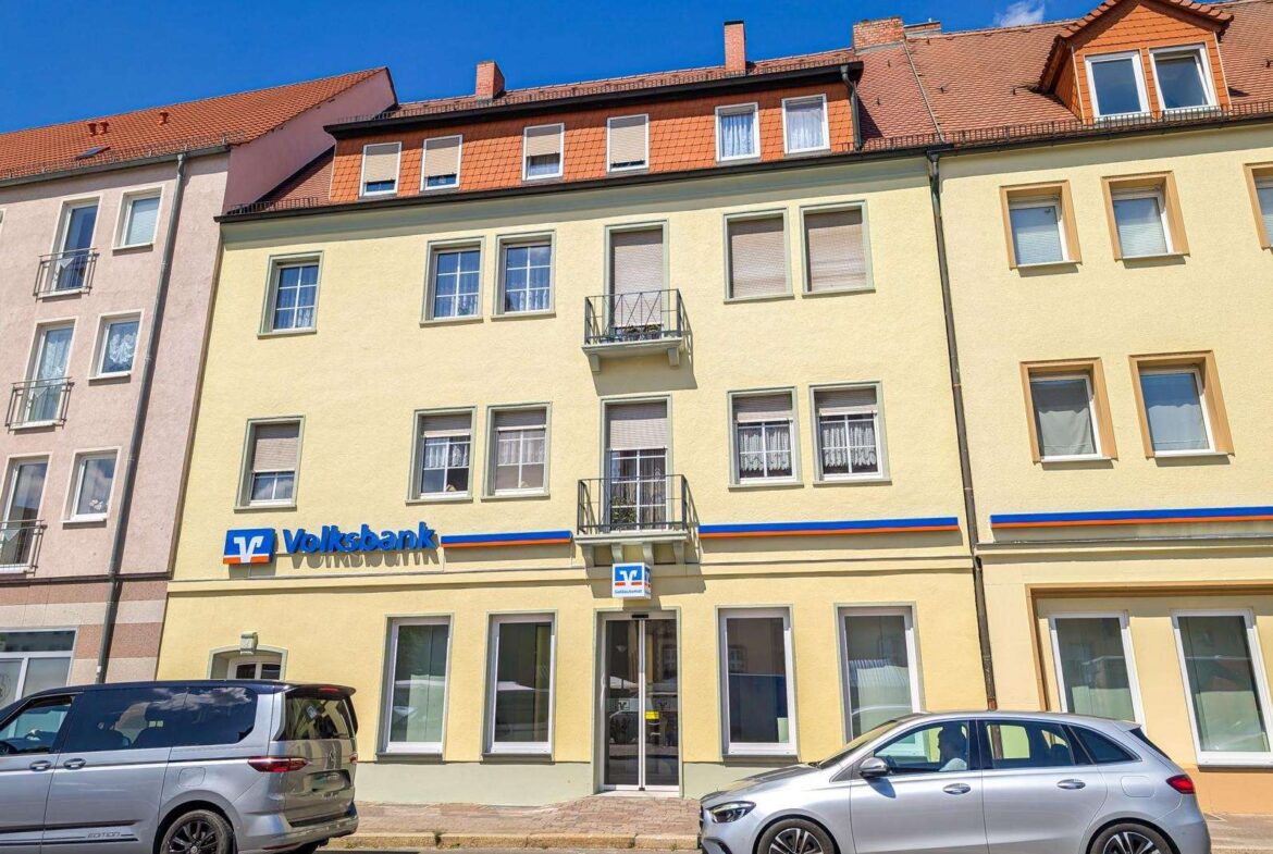 Yellow building with Volksbank sign on the ground floor, two balconies, and parked cars in front.