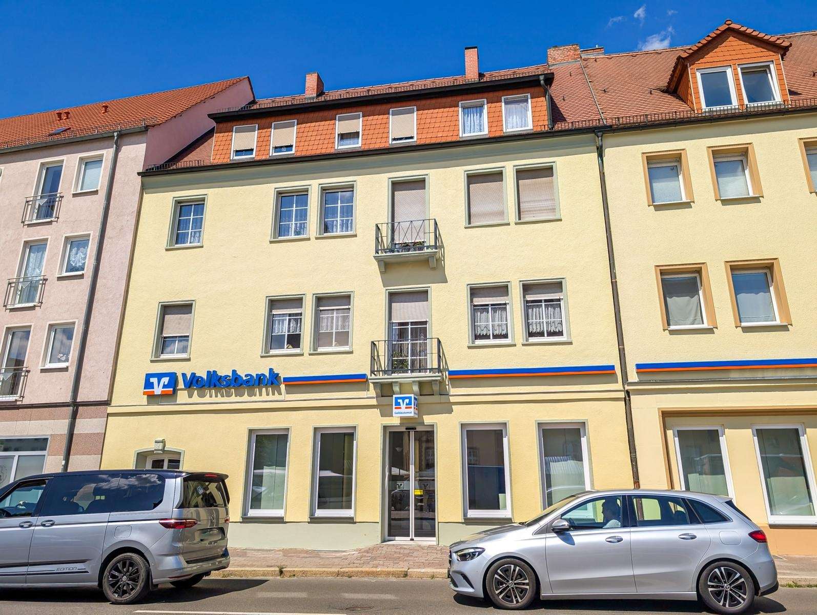 Yellow building with Volksbank sign on the ground floor, two balconies, and parked cars in front.