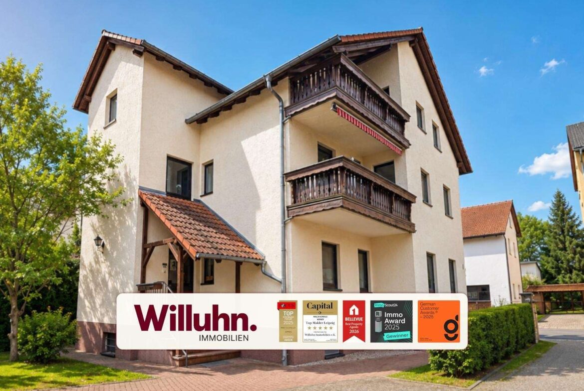 Three-story beige building with dark wooden balconies and a red-tiled roof; a Willuhn Immobilien banner is in the foreground.