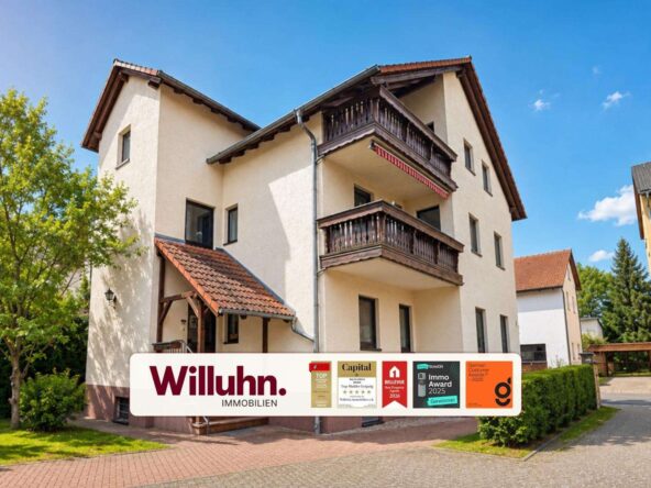 Three-story beige building with dark wooden balconies and a red-tiled roof; a Willuhn Immobilien banner is in the foreground.