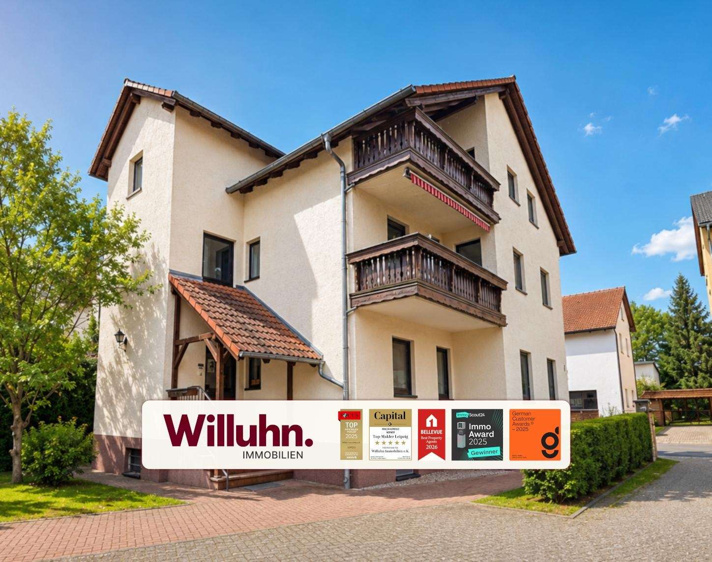 Three-story beige building with dark wooden balconies and a red-tiled roof; a Willuhn Immobilien banner is in the foreground.