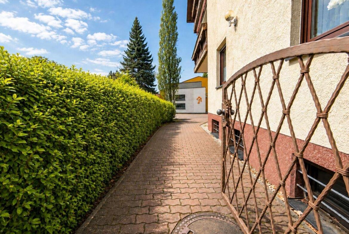 Narrow brick walkway flanked by a tall green hedge on the left and a beige building with a wrought-iron lattice gate on the right, under a blue sky with trees in the background.