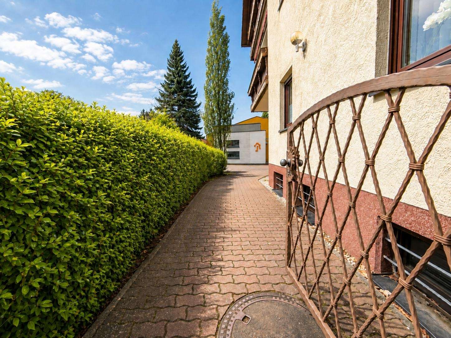 Narrow brick walkway flanked by a tall green hedge on the left and a beige building with a wrought-iron lattice gate on the right, under a blue sky with trees in the background.