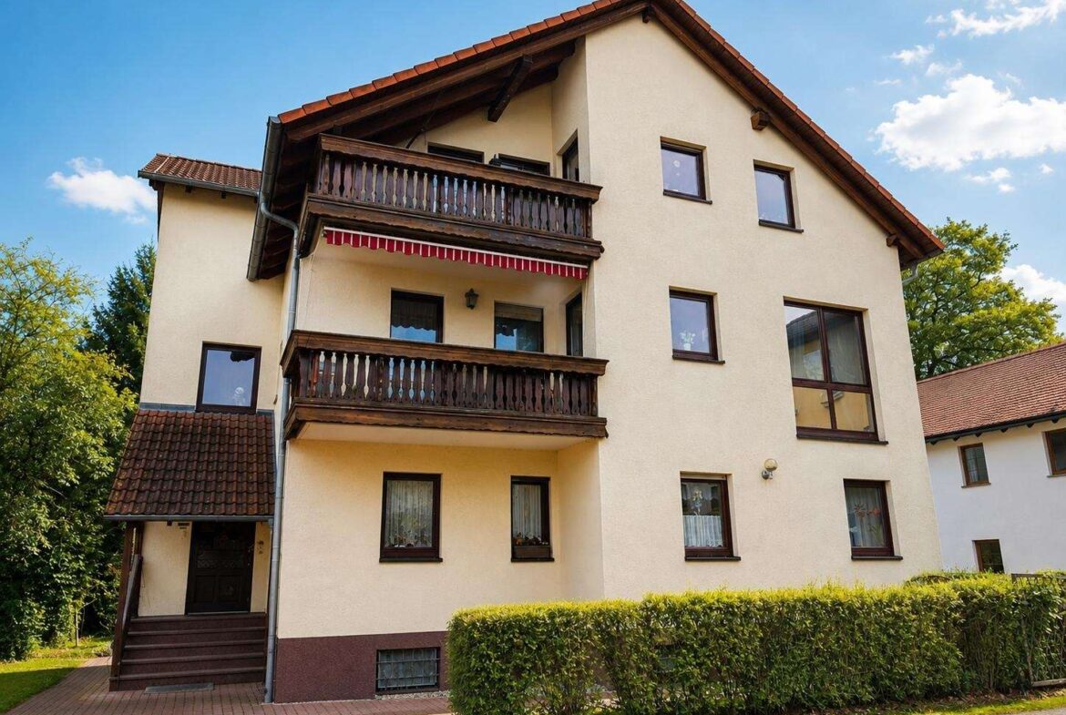 Three-story beige apartment building with dark wooden balconies and a red tile roof, seen behind a trimmed hedge under a blue sky.