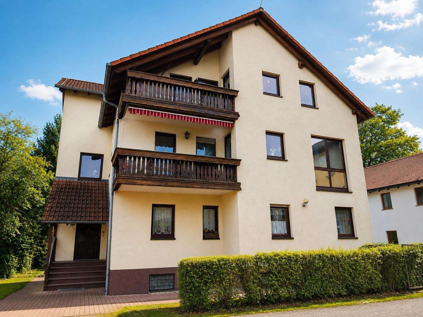Three-story beige apartment building with dark wooden balconies and a red tile roof, seen behind a trimmed hedge under a blue sky.