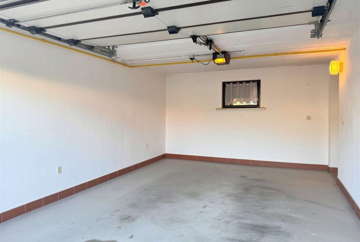 Empty garage interior with exposed overhead door hardware, a small window with sheer curtains, and a yellow gas pipe along the ceiling.