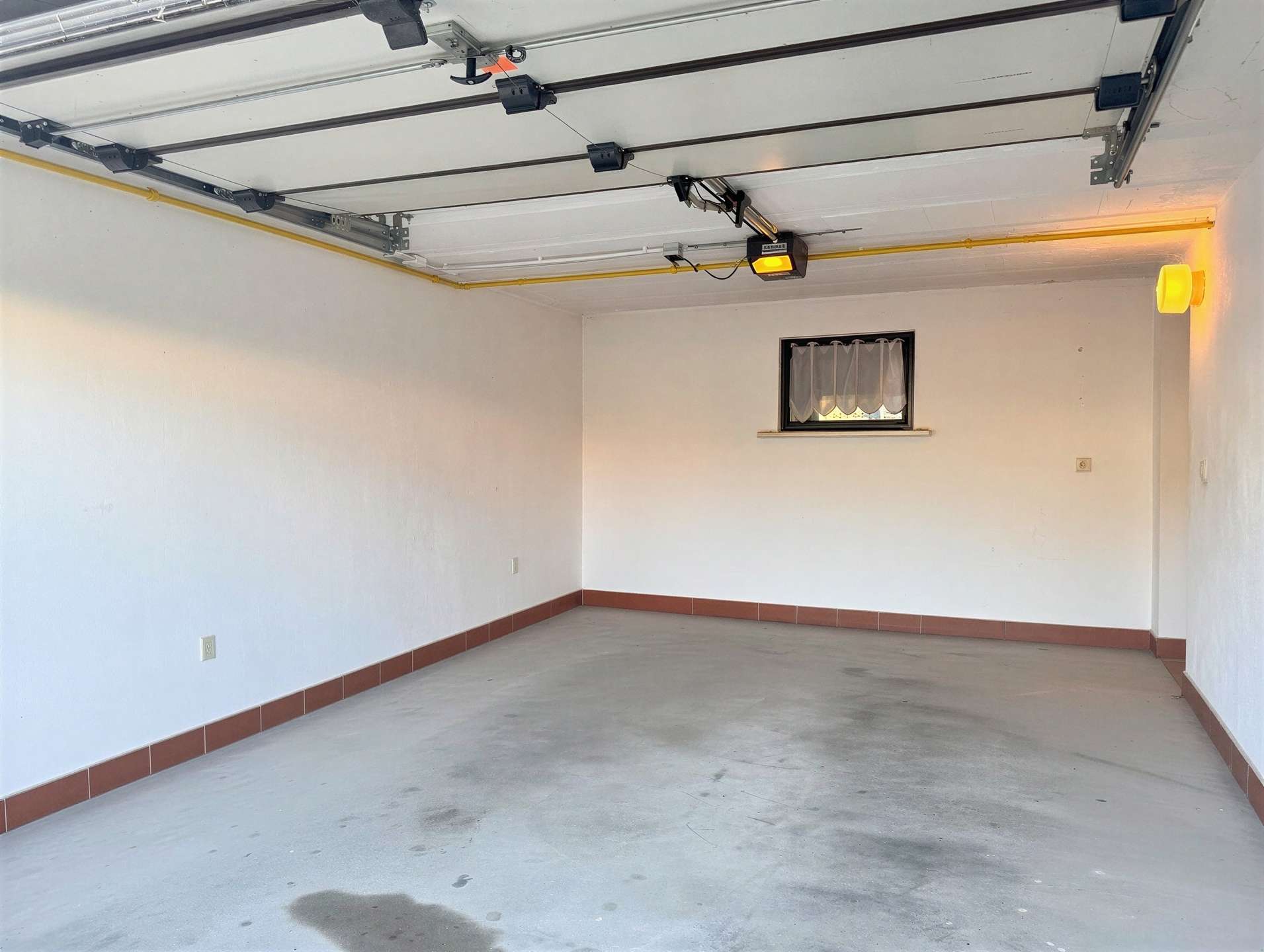 Empty garage interior with exposed overhead door hardware, a small window with sheer curtains, and a yellow gas pipe along the ceiling.