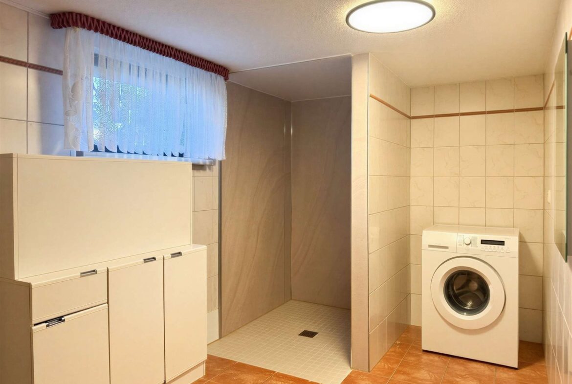Laundry room with a front-loading washing machine, white cabinets, and beige tiled walls; a small shower area sits to the left under a lace-curtained window.