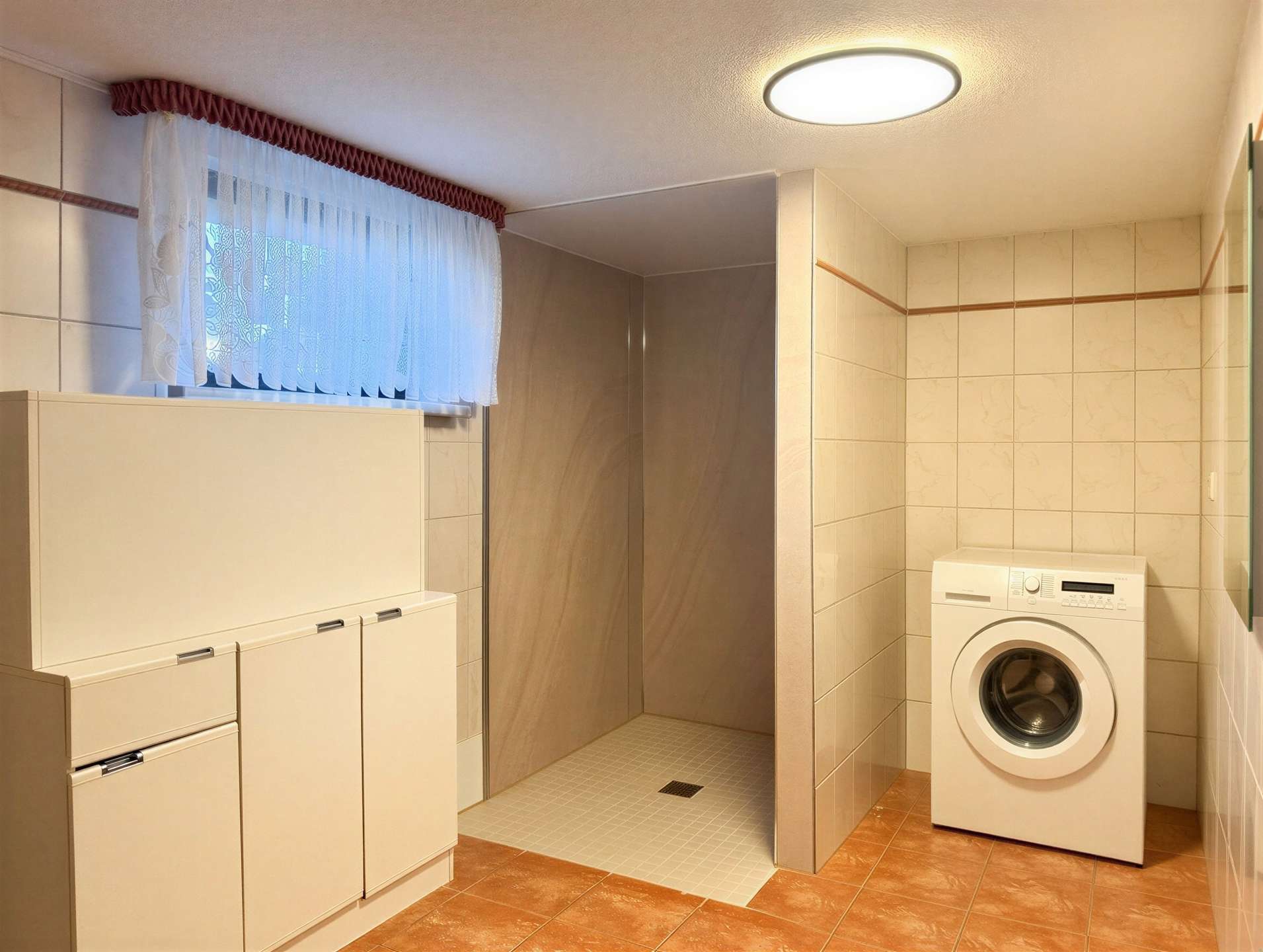 Laundry room with a front-loading washing machine, white cabinets, and beige tiled walls; a small shower area sits to the left under a lace-curtained window.