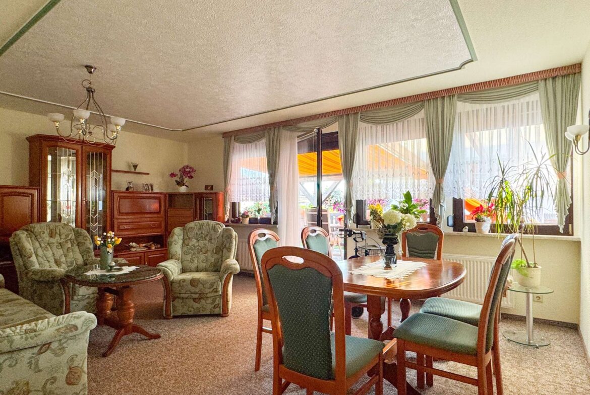 Cozy living and dining room with round wooden table, green upholstered chairs, and a chandelier, near large windows with sheer curtains.