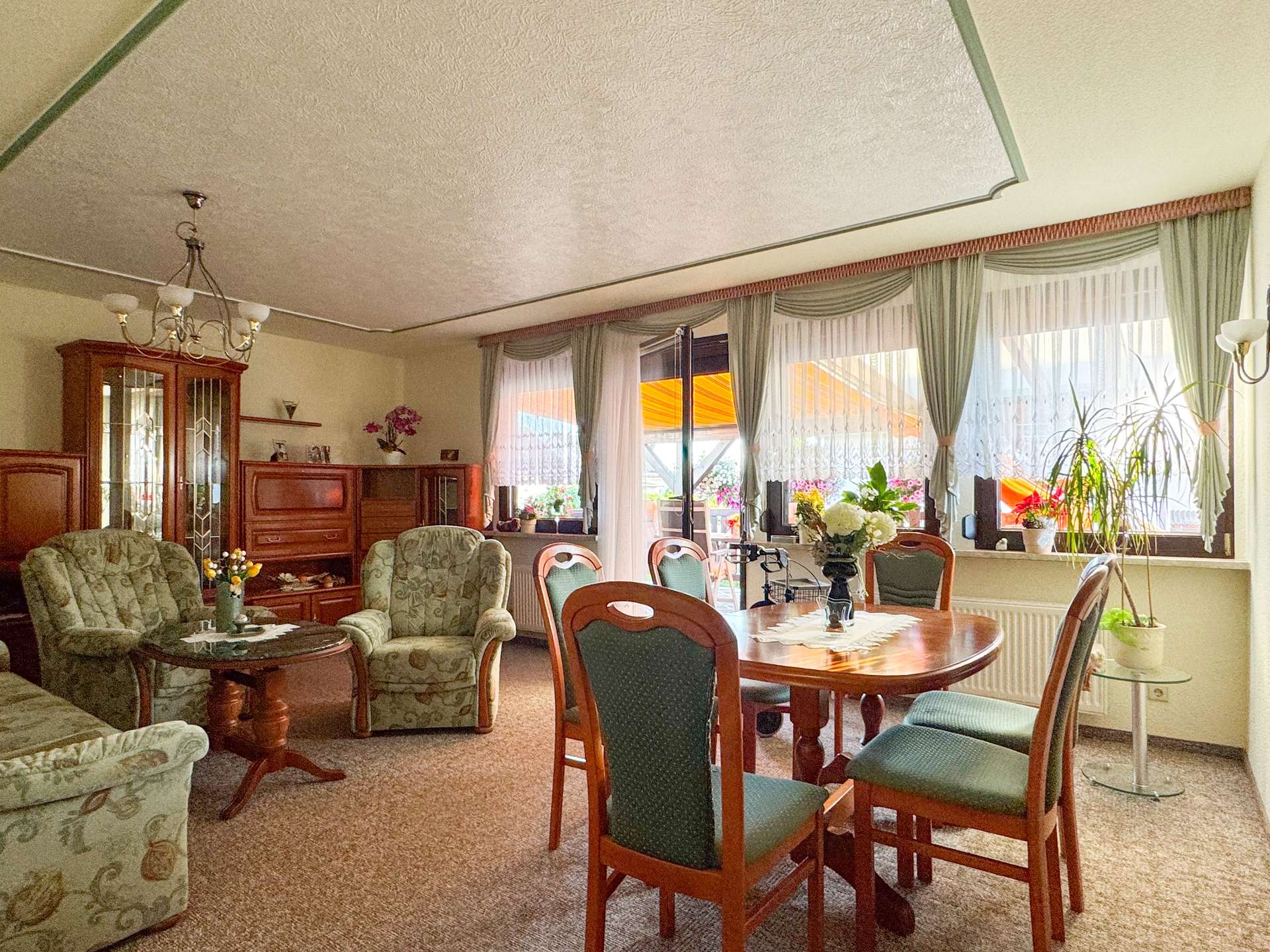 Cozy living and dining room with round wooden table, green upholstered chairs, and a chandelier, near large windows with sheer curtains.