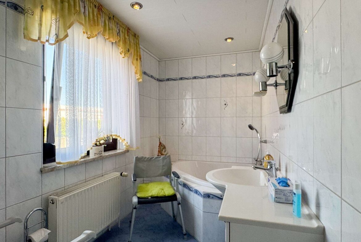 Small bright bathroom with white tiled walls, a bathtub, sink, and a folding chair by the window with sheer white and yellow curtains.