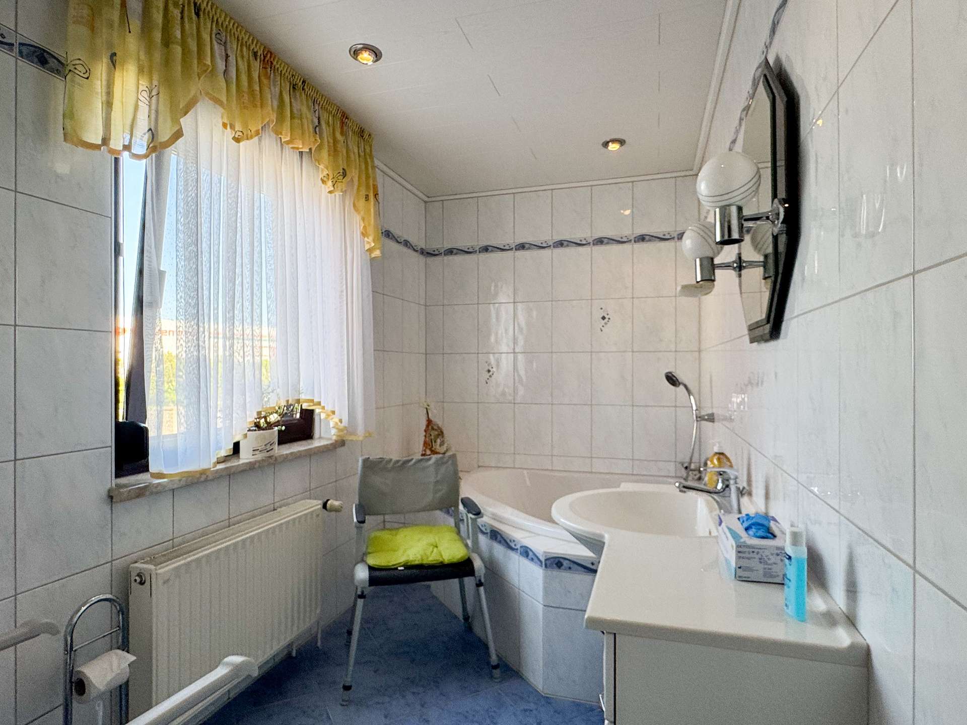 Small bright bathroom with white tiled walls, a bathtub, sink, and a folding chair by the window with sheer white and yellow curtains.