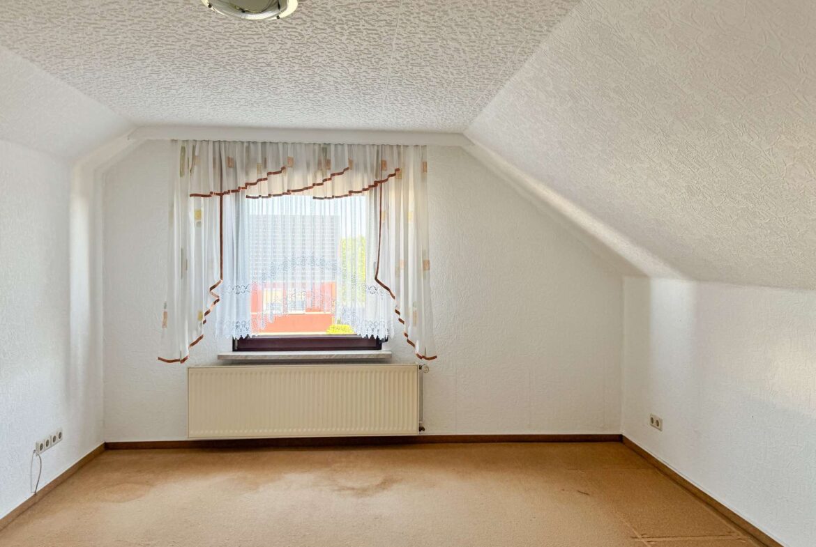 Small empty room with a window, sheer curtains, and a radiator beneath the window on a beige carpeted floor.