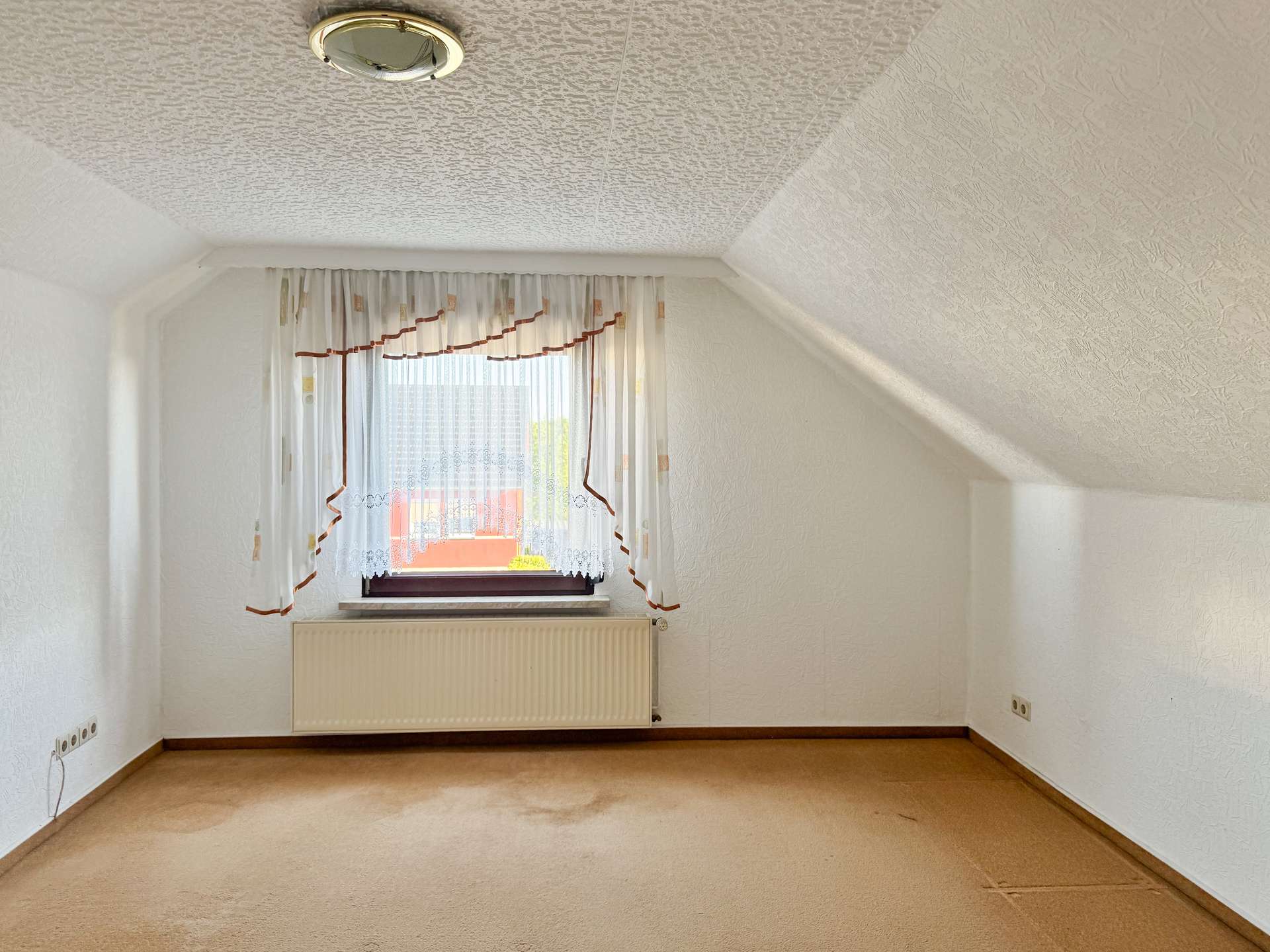 Small empty room with a window, sheer curtains, and a radiator beneath the window on a beige carpeted floor.